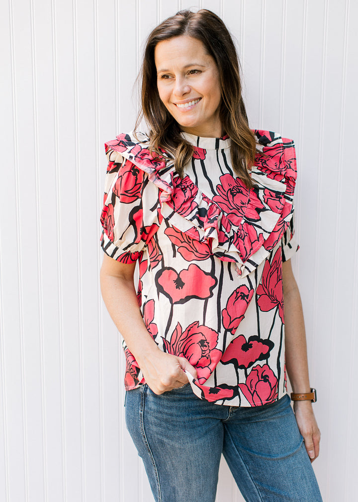 Model wearing a cream top with rose colored flowers, a bib ruffle detail and short sleeves.