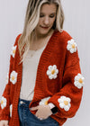Model wearing a rust cardigan with white and yellow embroidered daisies and long sleeves. 
