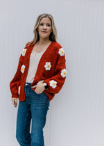 Model wearing jeans and a rust cardigan with white and yellow embroidered daisies and long sleeves. 