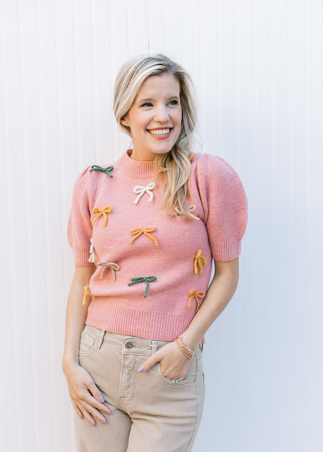 Model wearing khakis with a dusty pink sweater with cream, taupe and green bows and short sleeves.