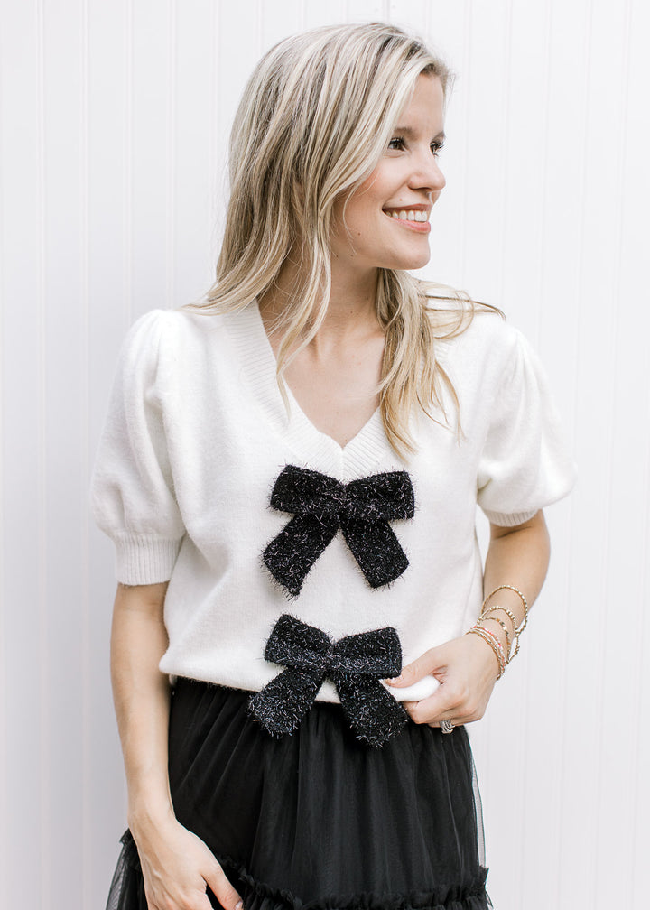 Model wearing a cream v-neck sweater with 2 black shimmer bows and short sleeves.