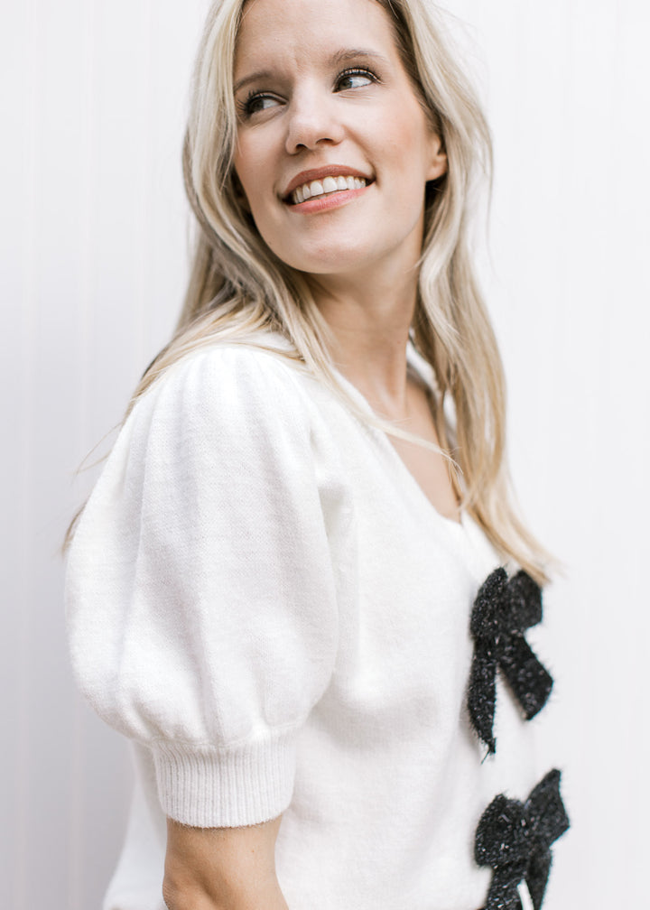 Close up of short puff sleeves and v-neck on a cream top with two black shimmer bows.