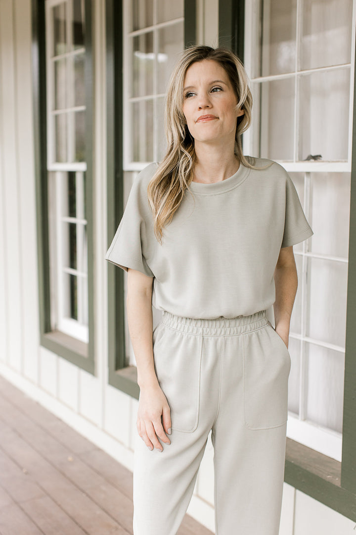 Model wearing a sage jumpsuit with an elastic waist, short sleeves and pockets.