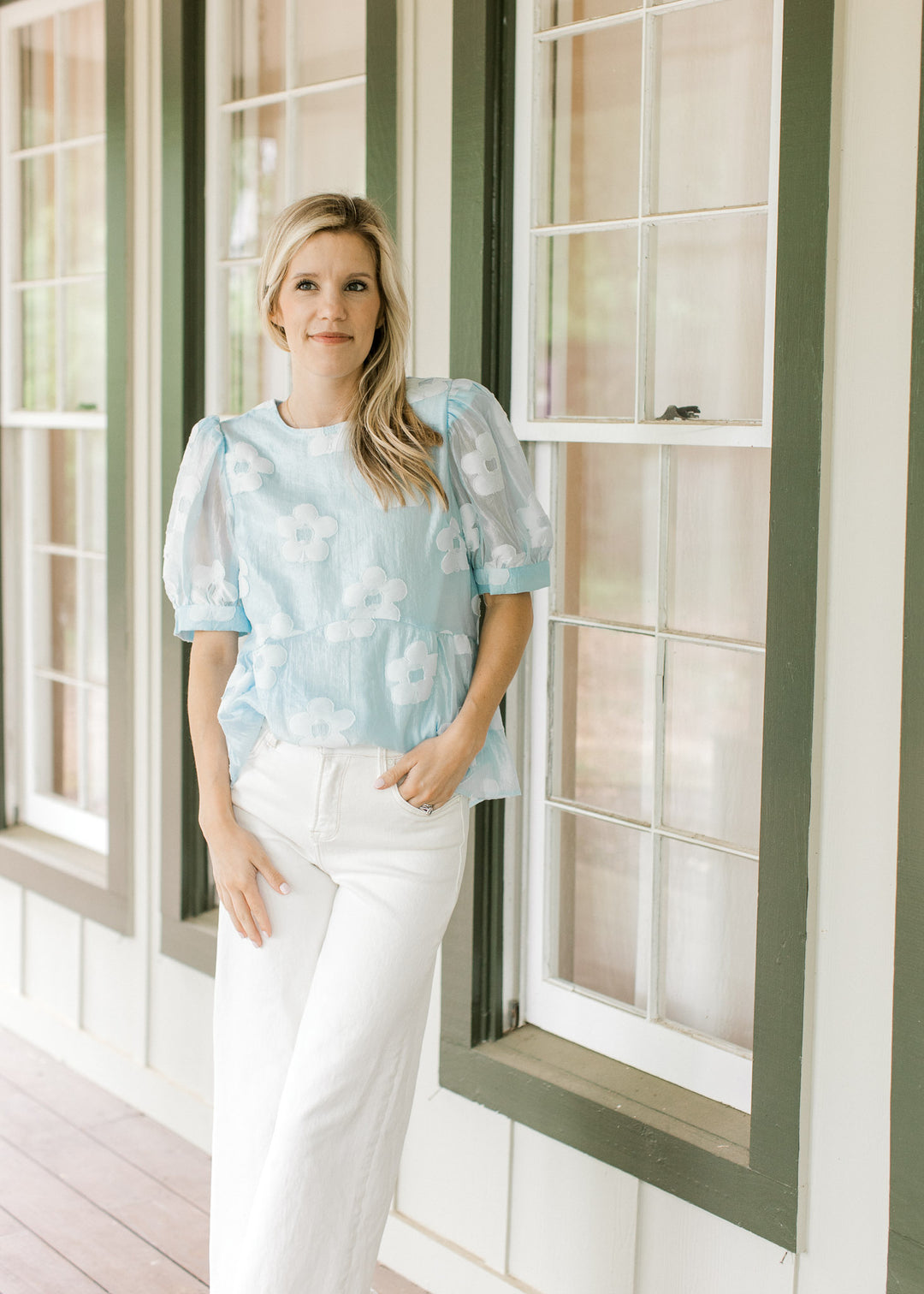 Model wearing white jeans with a light blue top with stitched flowers and sheer short sleeves.