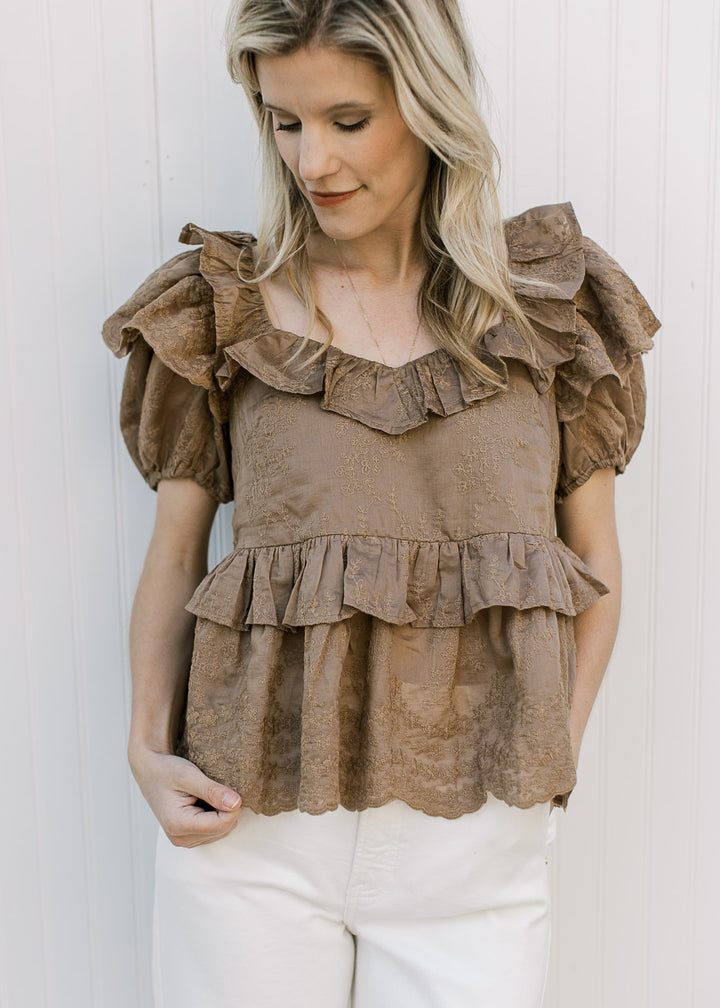 Model wearing a golden brown top with embroidered floral, short sleeves and ruffle details.