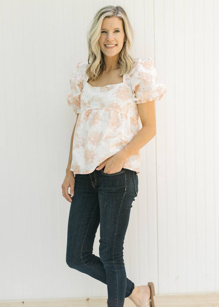 Model wearing jeans and sandals with a white and coral embroidered floral top with bubble sleeves.