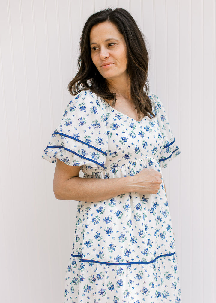 Model wearing a cream maxi with blue floral, navy piping, bubble short sleeves, v-neck and pockets.