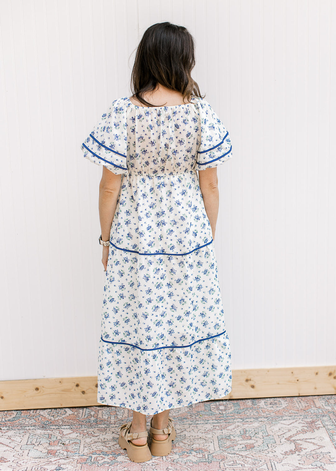 Back view of Model wearing a cream maxi with blue floral, bubble short sleeves, v-neck and pockets.