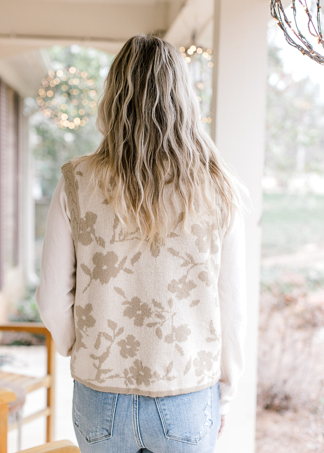 Back view of Model wearing a cream sleeveless cardigan with taupe flowers and edging.