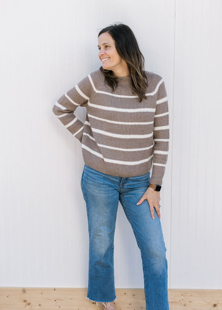 Model wearing a taupe sweater with white stripes, with long sleeves and a ribbed material.