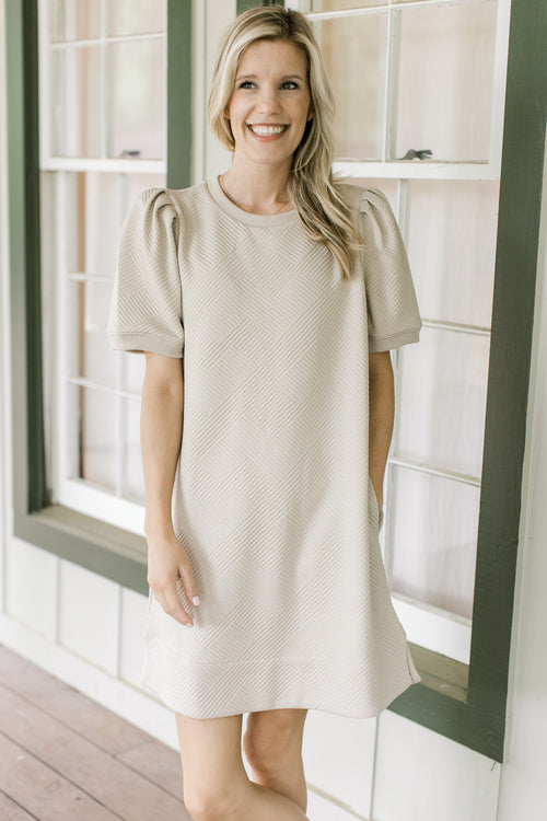 Model wearing a taupe dress with pleated shoulders, short sleeves and embossed pattern.