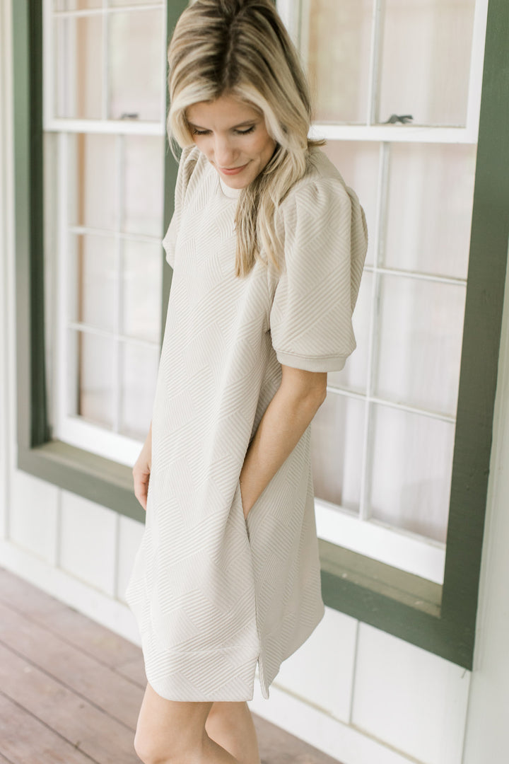 Model wearing a taupe dress with pleated shoulders, short sleeves and pockets.