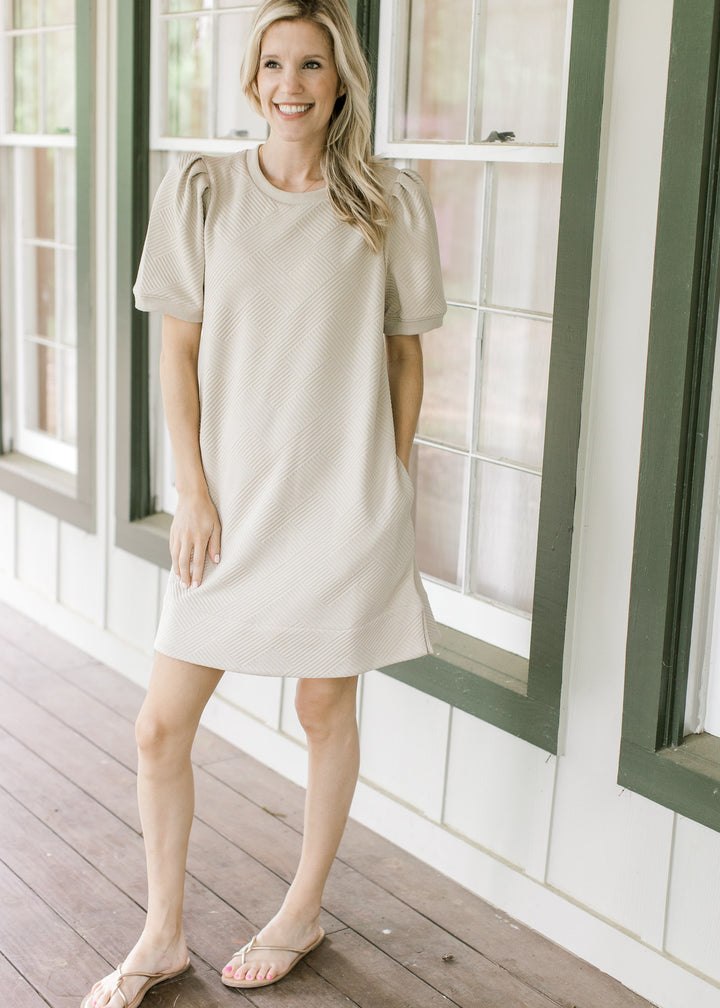 Model wearing sandals with a taupe dress with pleated shoulders, short sleeves and embossed pattern.