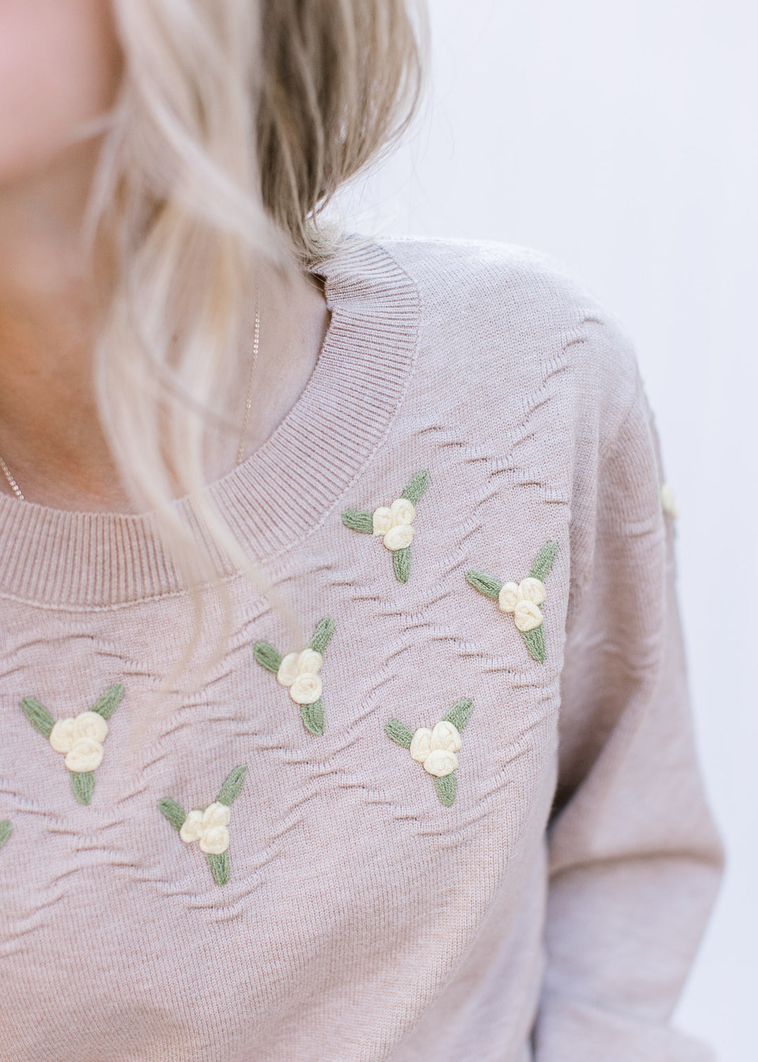 Model wearing a beige sweater with green and yellow flowers at neck, long sleeves and round neck.