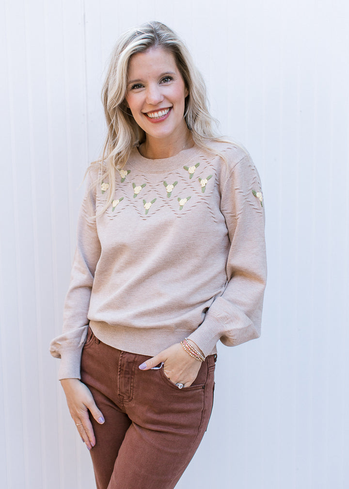 Model wearing a beige long sleeve sweater with green and yellow flowers at neck and a round neck.