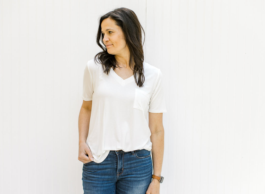 Model wearing a ivory colored short sleeve v-neck tee with a chest pocket and blue jeans.
