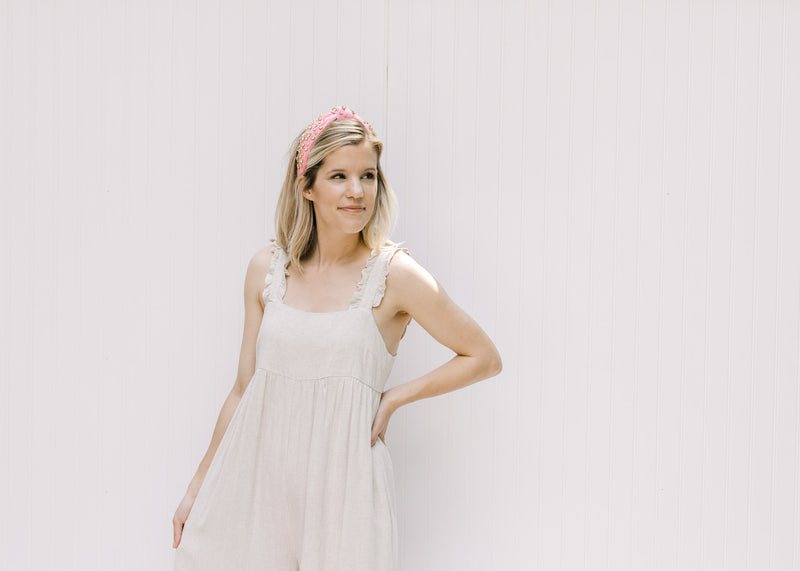 Model wearing a sand colored jumpsuit with a pink bejeweled headband with a knotted fabric design. 