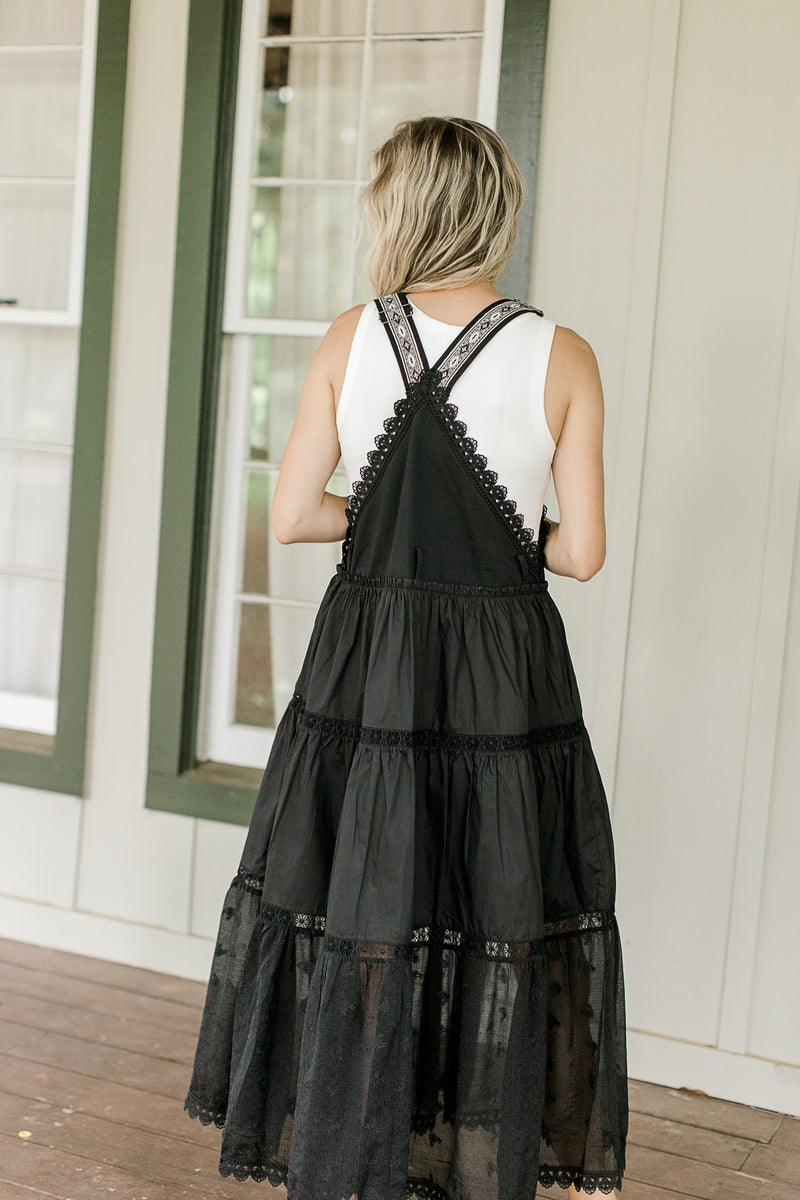 Back view of Model wearing a black overall maxi with adjustable straps and a square neck.