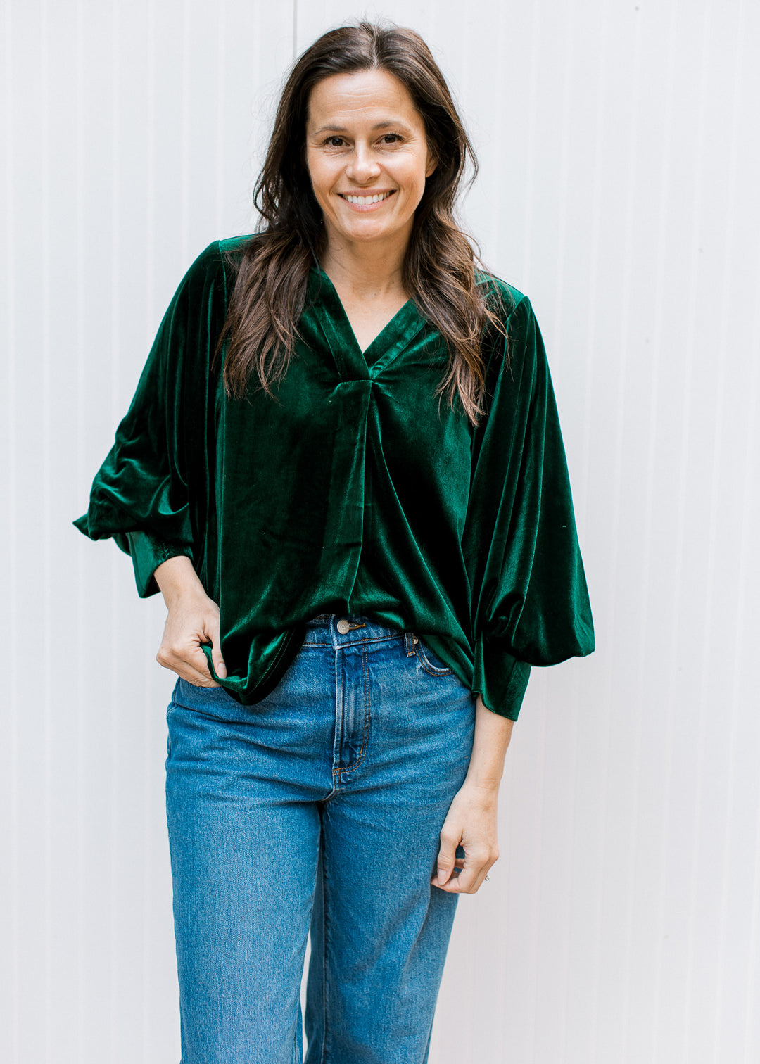 Model holding the hem of a bottle green velvet top with V-neckline and three-quarter bubble sleeves, worn with jeans.