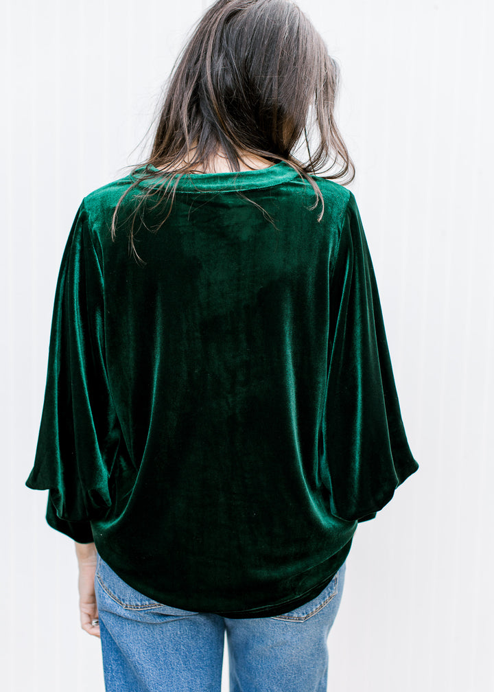 Back view of bottle green velvet top with rounded neckline, relaxed drape, bubble sleeves and curved hem over jeans.