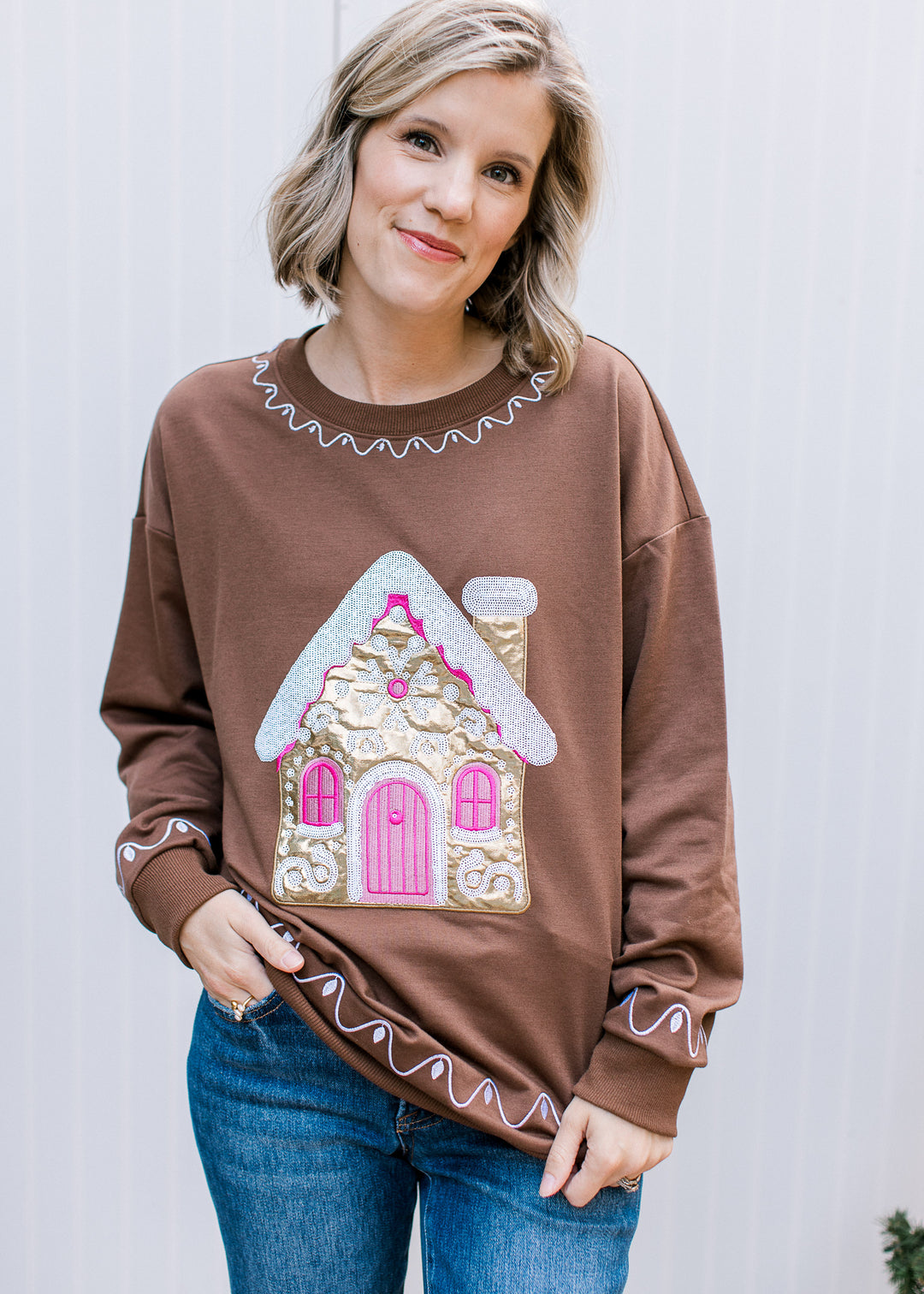 Brown Christmas pullover with gold gingerbread house, pink door and windows, white scalloped trim and long sleeves.