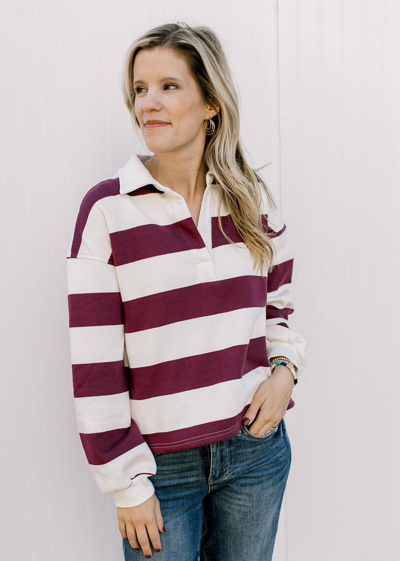 Model wearing a burgundy and cream rugby stripe long sleeve top with cuffs, v-neck and a collar.