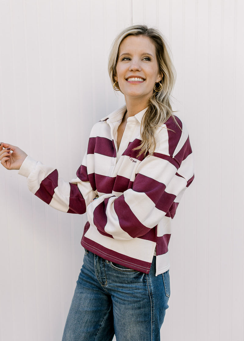 Model wearing jeans and a burgundy and cream rugby stripe long sleeve top with a v-neck and collar. 