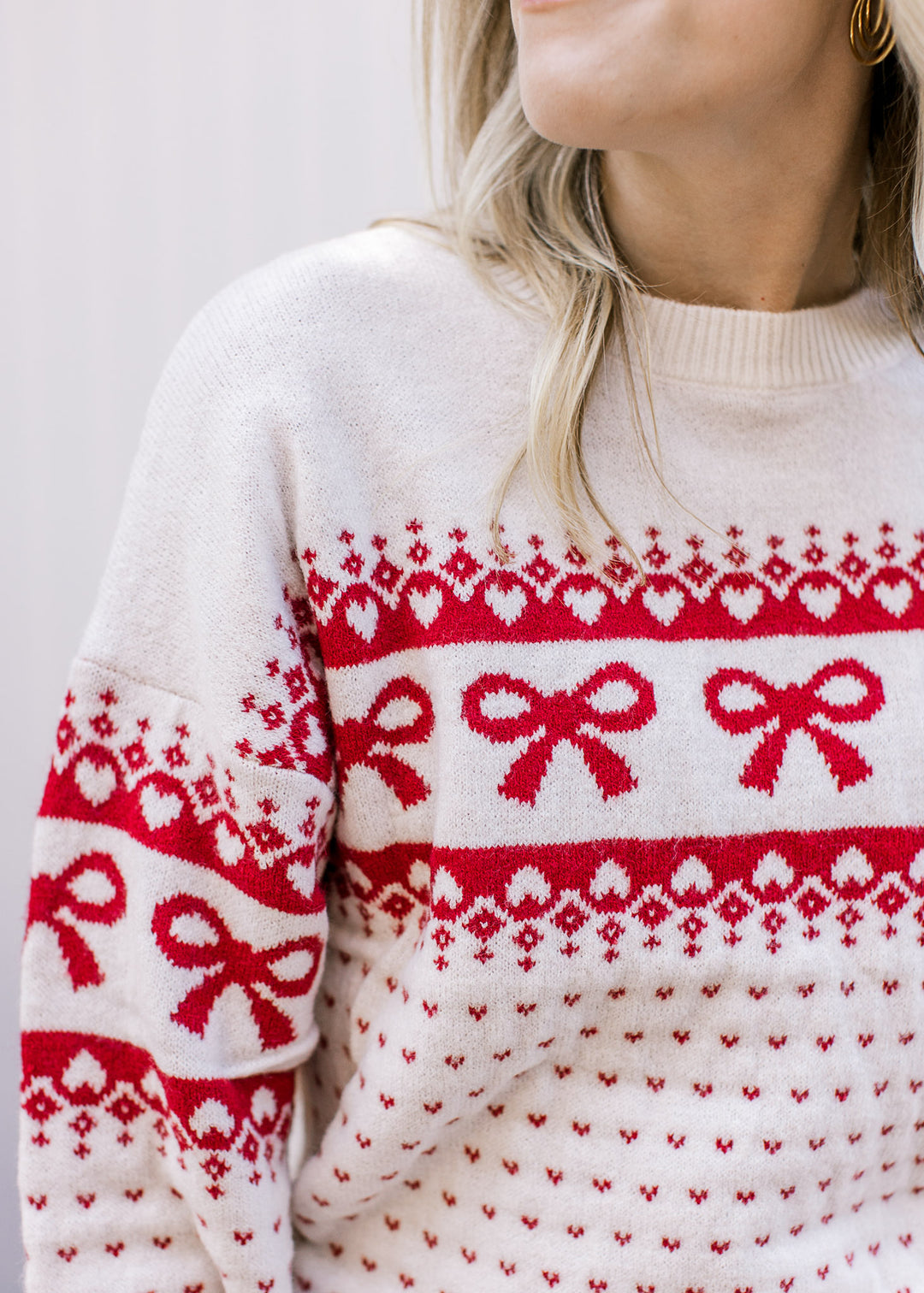 Close up of red bows and heart accents on a cream sweater with a round neck and long sleeves. 