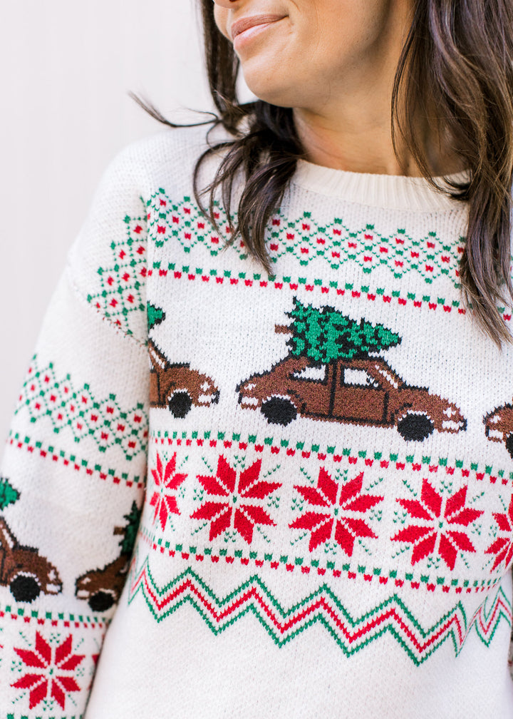 Model wearing a cream sweater with red and green accents and brown trucks carrying a Christmas tree.