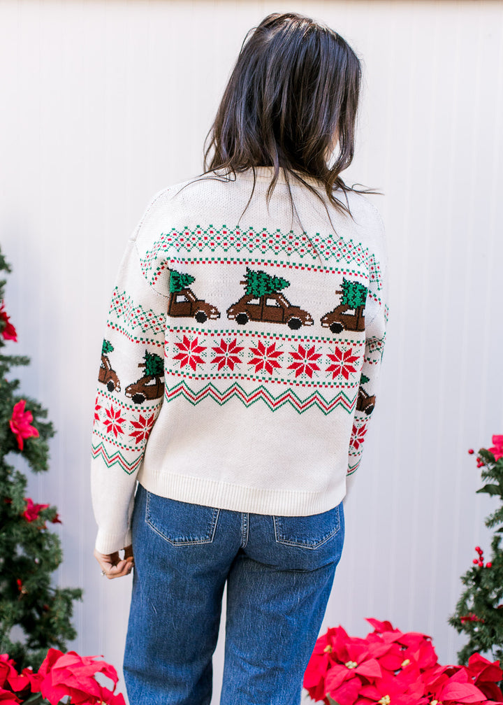 Back view of Model wearing a cream long sleeve sweater with brown trucks carrying a Christmas tree.