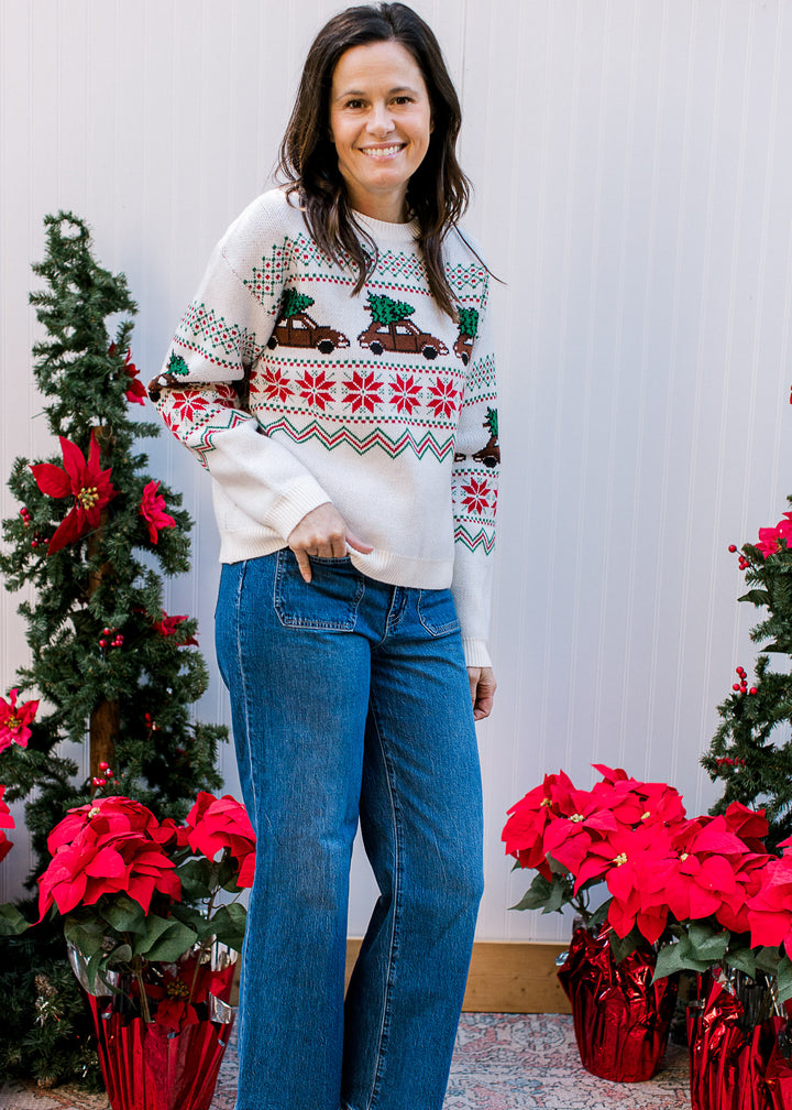 Model wearing a cream long sleeve sweater with brown trucks carrying a Christmas tree.