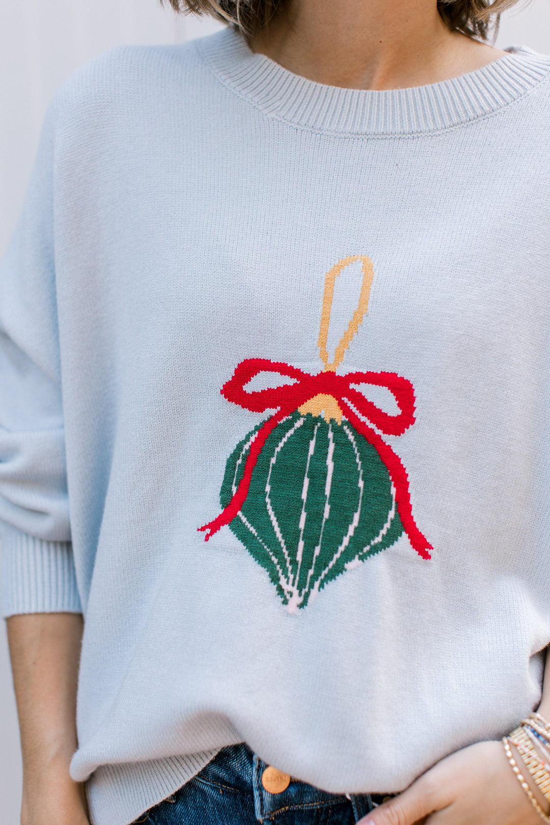 Close-up of pale blue sweater showing green Christmas ornament with white stripes, red bow and gold hanging loop.