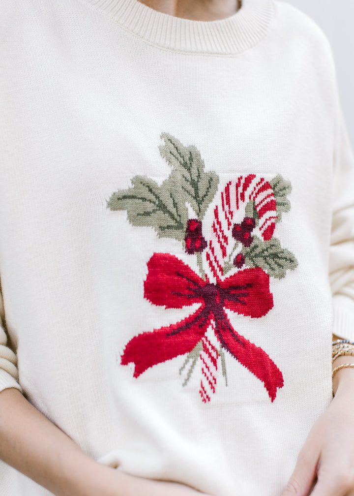 Close-up of cream Christmas sweater showing red-and-white candy cane, green holly leaves, red berries and a red bow.
