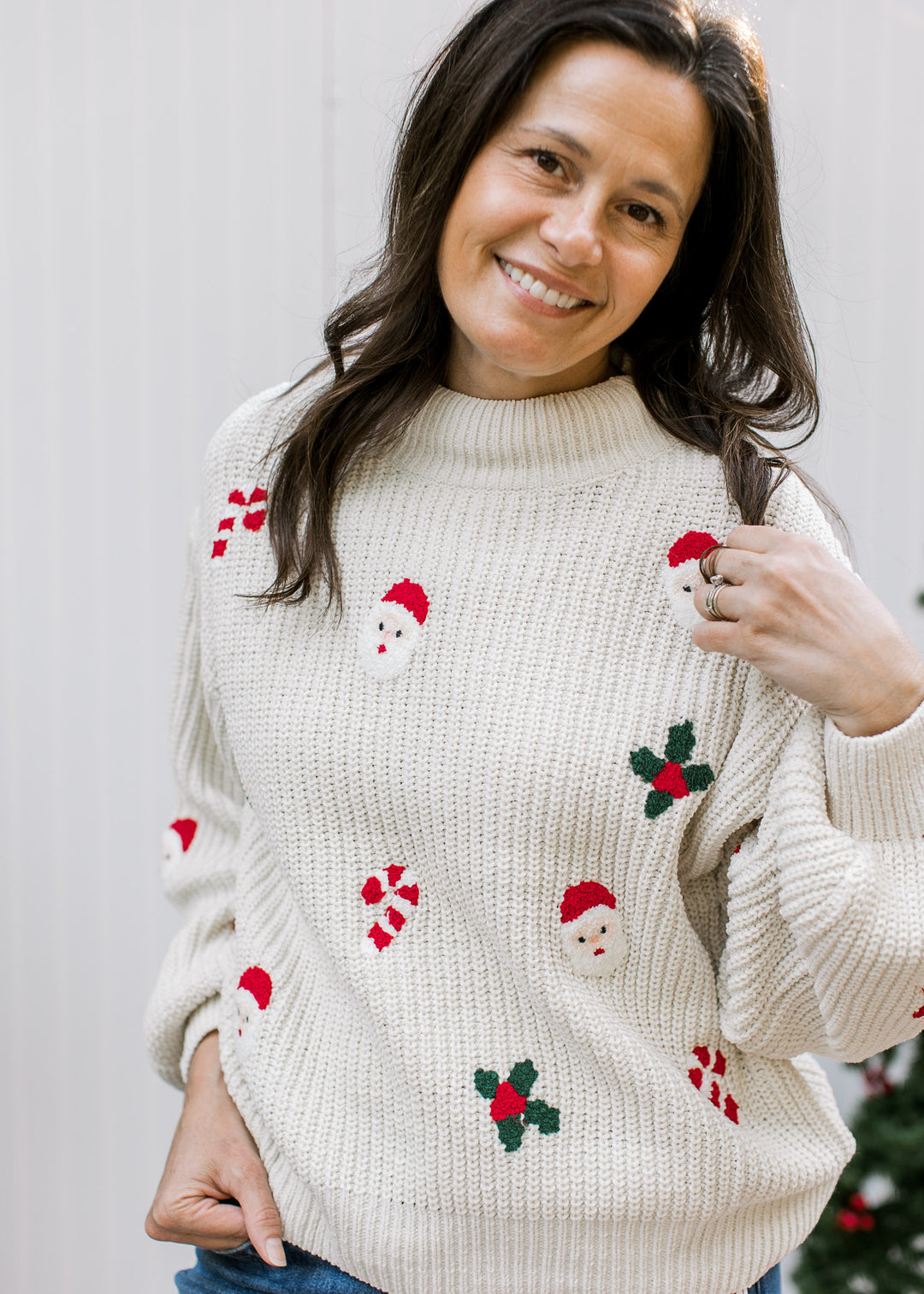 Model holding the shoulder of a cream Christmas sweater with Santa faces, red candy canes and green holly motifs.