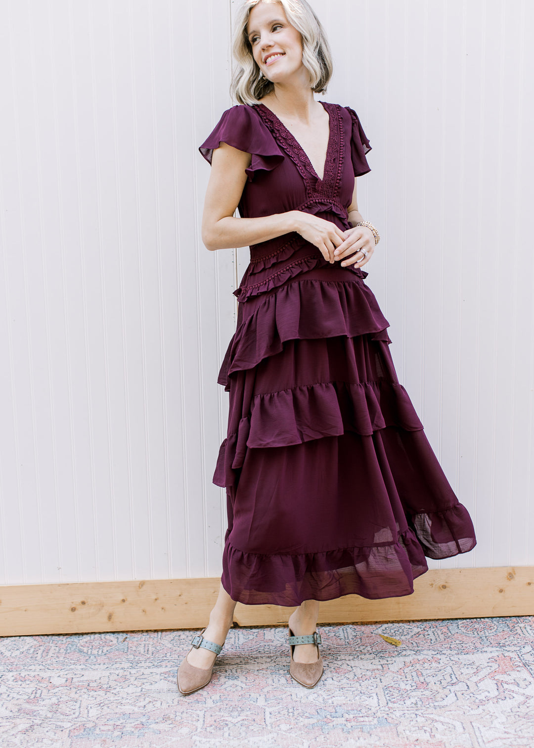 Angled view of dark wine dress with lace V-neckline and cascading skirt ruffles, styled with buckled heels.