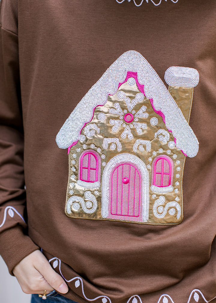 Close-up of gold gingerbread house on brown pullover, showing white sequin roof, pink windows and snowflake detail.
