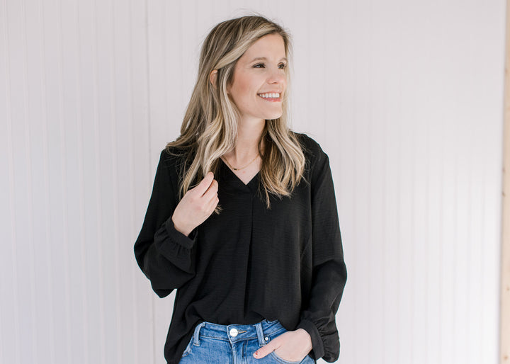 Model wearing a black v-neck top with long sleeves tucked into jeans. 