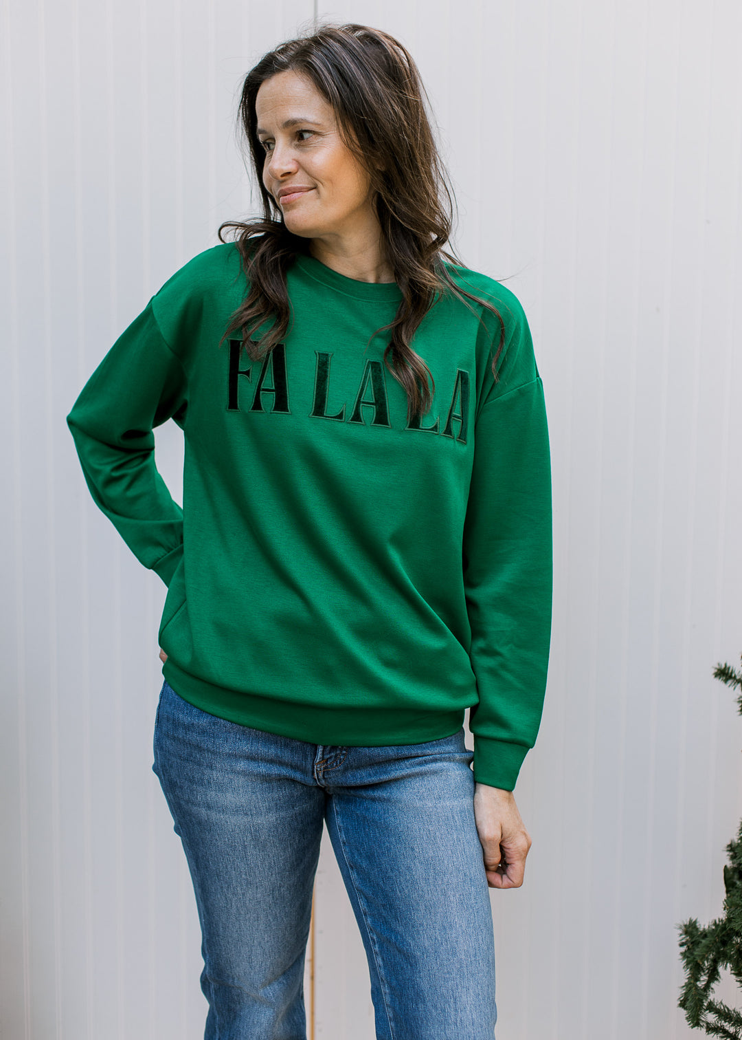 Front view of green FA LA LA pullover with velvet lettering, long sleeves and banded hem, styled with blue jeans.
