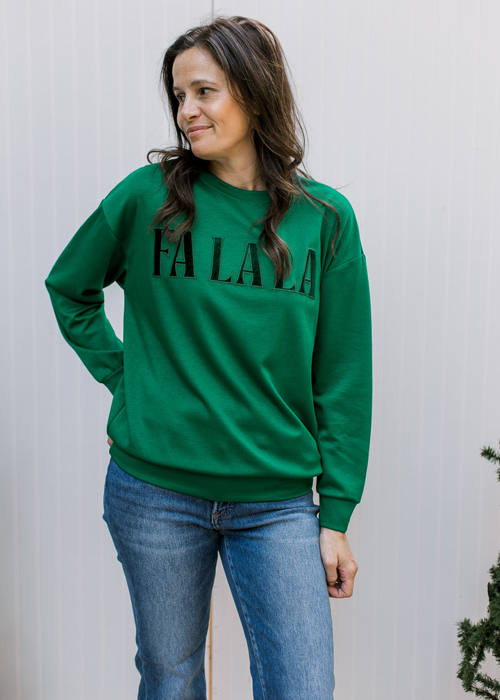 Front view of green FA LA LA pullover with velvet lettering, long sleeves and banded hem, styled with blue jeans.
