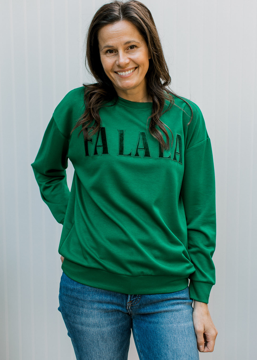  Model wearing green velvet patch FA LA LA pullover with relaxed drape, round neck and cuffed sleeves over jeans.