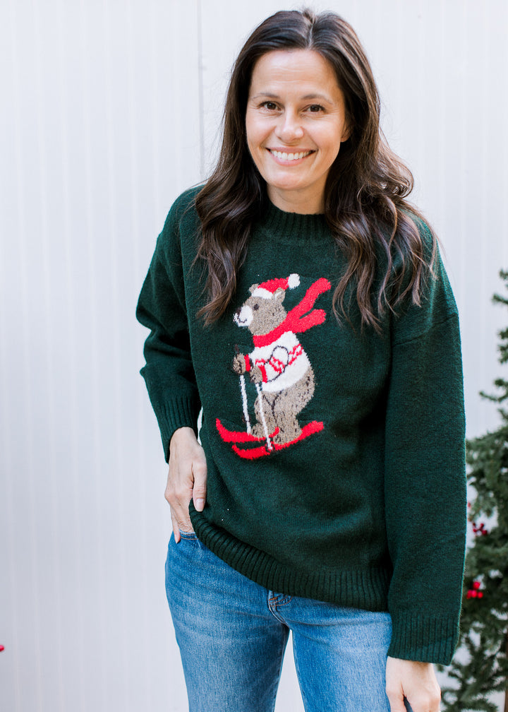 Model wearing hunter green Skiing Bear Sweater with red ski graphic, ribbed crew neck and cuffs, styled with jeans.