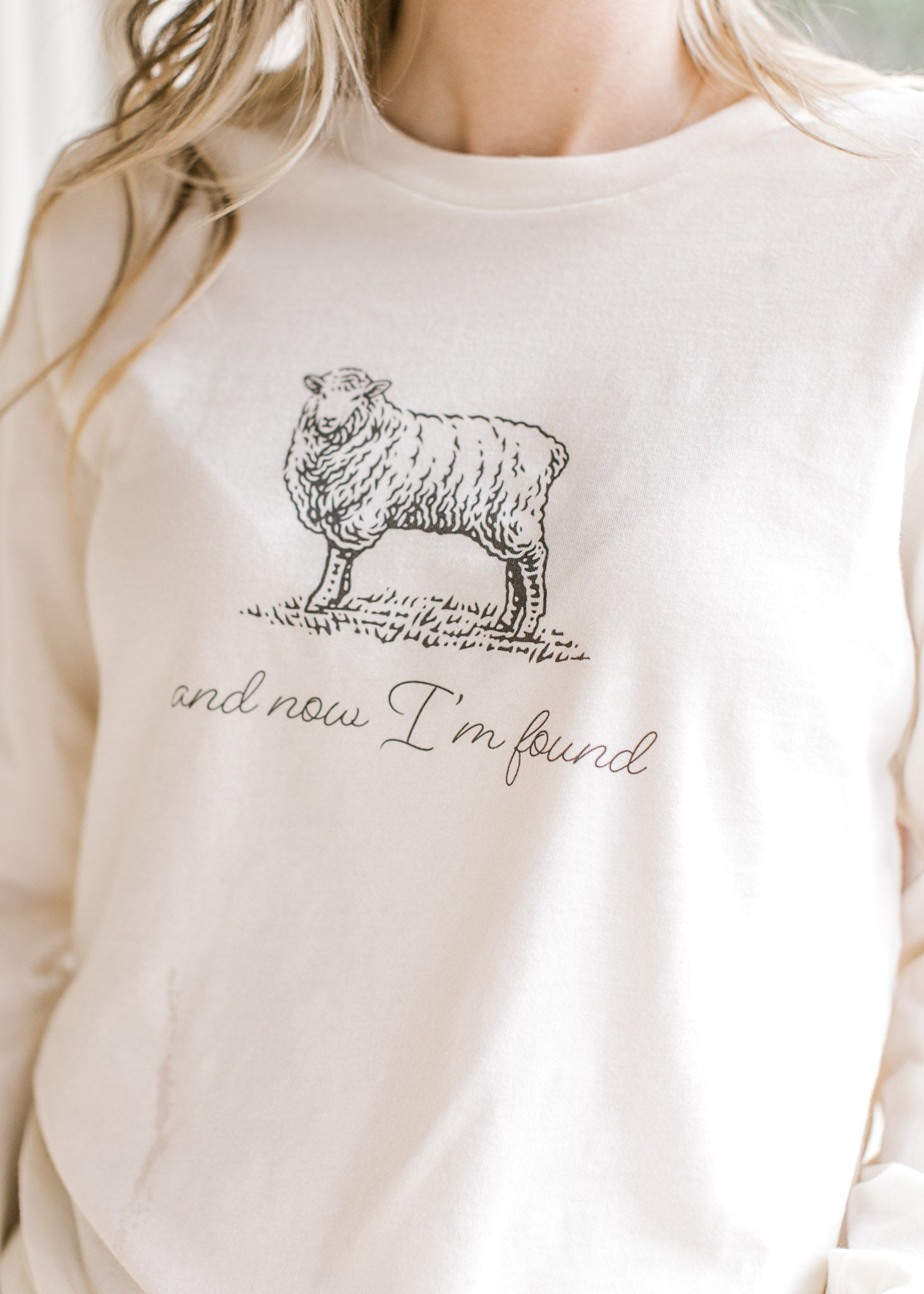 Model wearing a cream long sleeve tee with script “and now I’m found” and black sheep illustration.