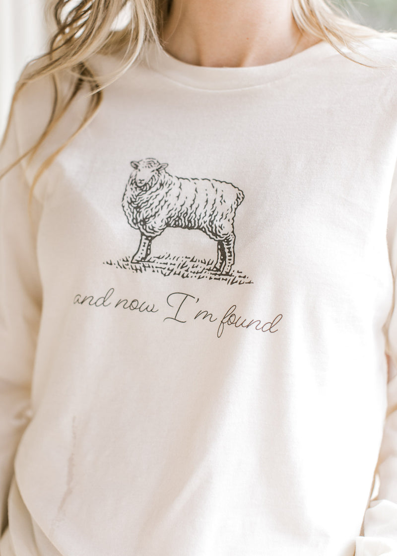 Model wearing a cream long sleeve tee with script “and now I’m found” and black sheep illustration.