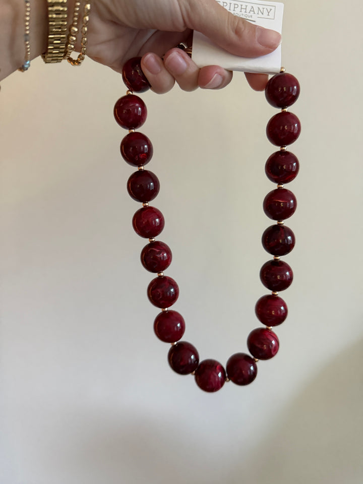 Hand holding a maroon beaded bubble necklace, showing its rounded burgundy beads, glossy finish and gold-tone spacers.