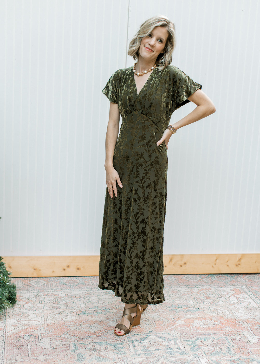 Model wearing olive velvet floral maxi dress with V-neckline and cap sleeves, styled with brown wedge sandals.