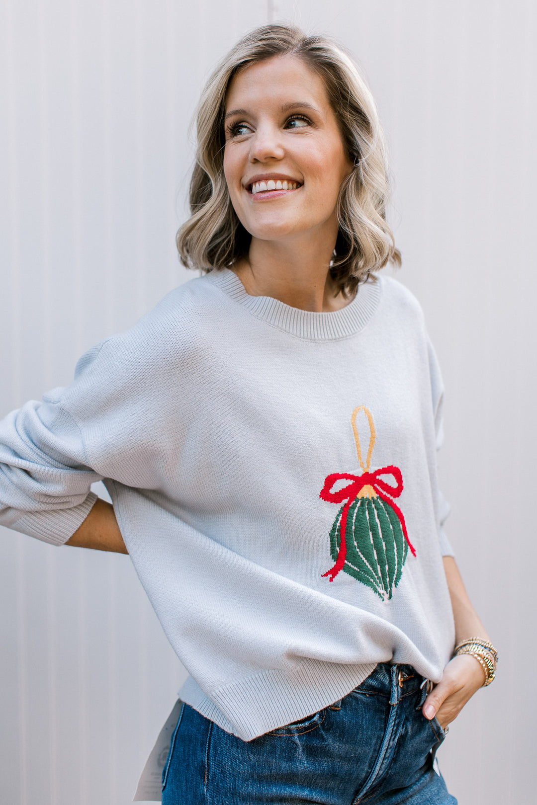 Pale blue Christmas sweater with a green ornament and red bow, ribbed crew neck and relaxed fit, styled with jeans.