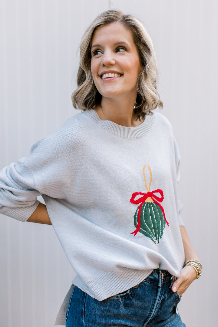 Pale blue Christmas sweater with a green ornament and red bow, ribbed crew neck and relaxed fit, styled with jeans.