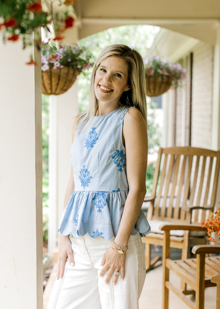 Model wearing white pants and a baby blue top with embroidered deep blue floral and scalloped hem.
