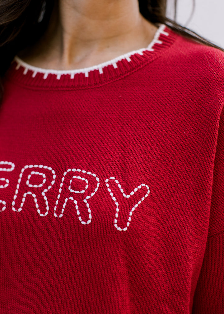 Close-up of red Christmas sweater showing cream stitched MERRY lettering and blanket stitching along ribbed crew neck.