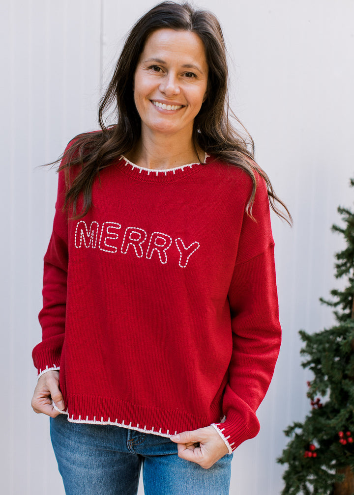 Model holding the hem of red MERRY sweater, showing cream outlined letters and contrast stitching at cuffs and hem.